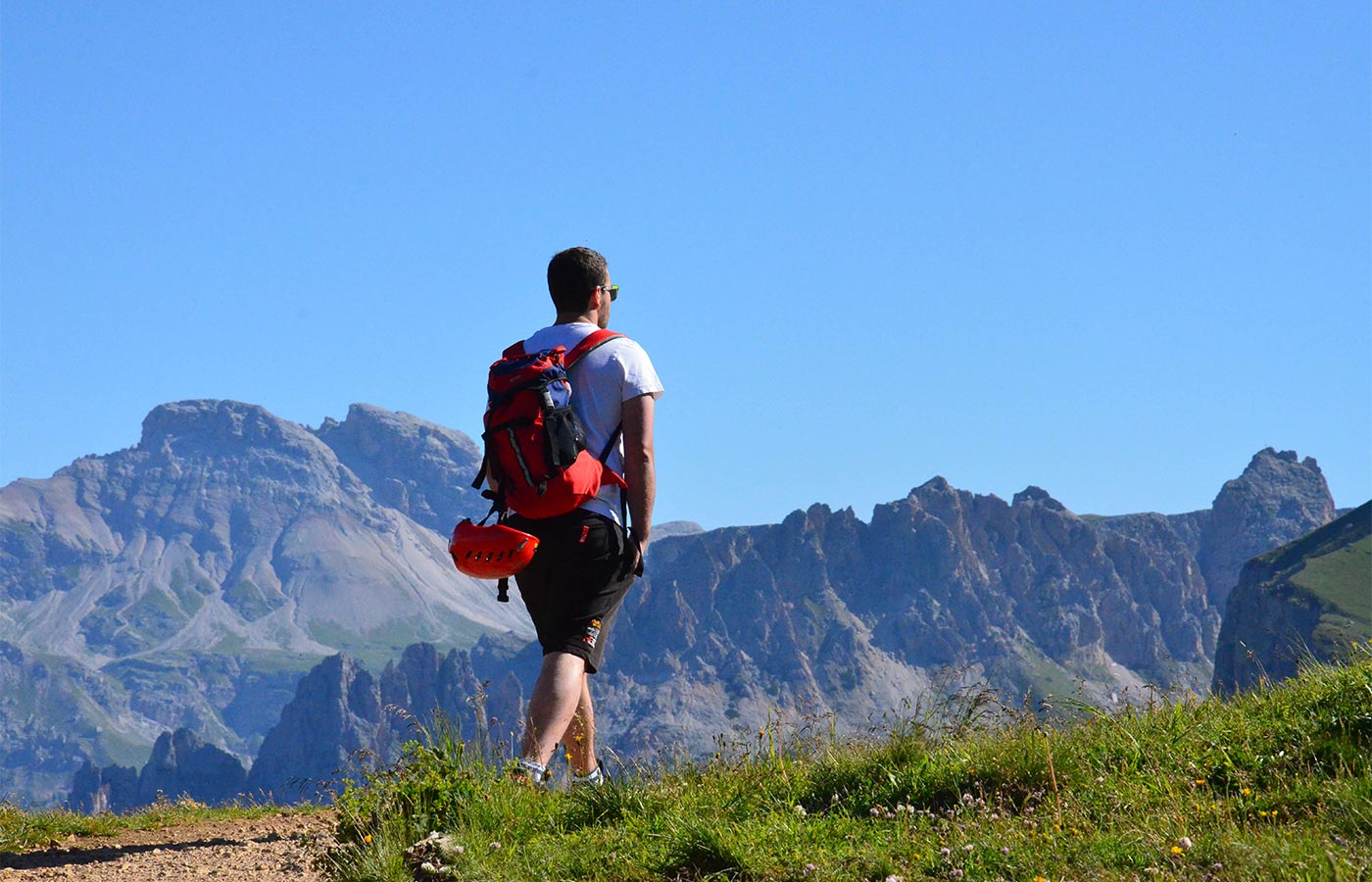 ein Wanderer mit Sonnenbrillen in kurzärmligem T-shirt und kurzen Hosen mit rotem Rucksack an dem ein roter Helm angebunden ist auf dem Rücken, beim Wandern in Wolkenstein
